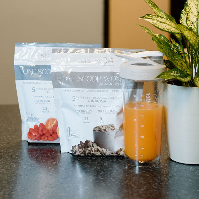 Two stand-up pouches of One Scoop Protein powder sit on a dark countertop beside a clear shaker bottle filled with orange liquid and a potted green plant.