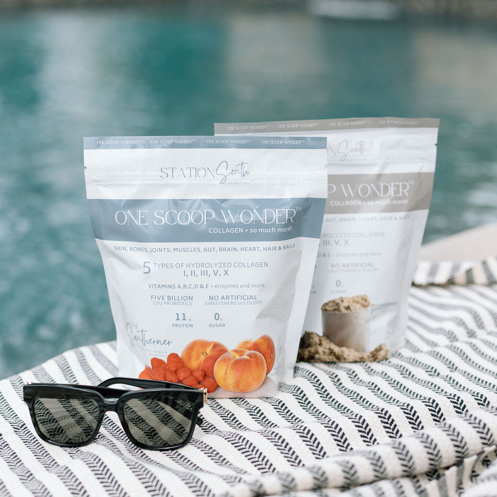 Two stand-up pouches of Station Q One Scoop Wonder Whole Body Wellness Blend powder are displayed on a striped blanket next to a swimming pool, with black sunglasses resting in front. The background shows a blurred outdoor poolside setting.