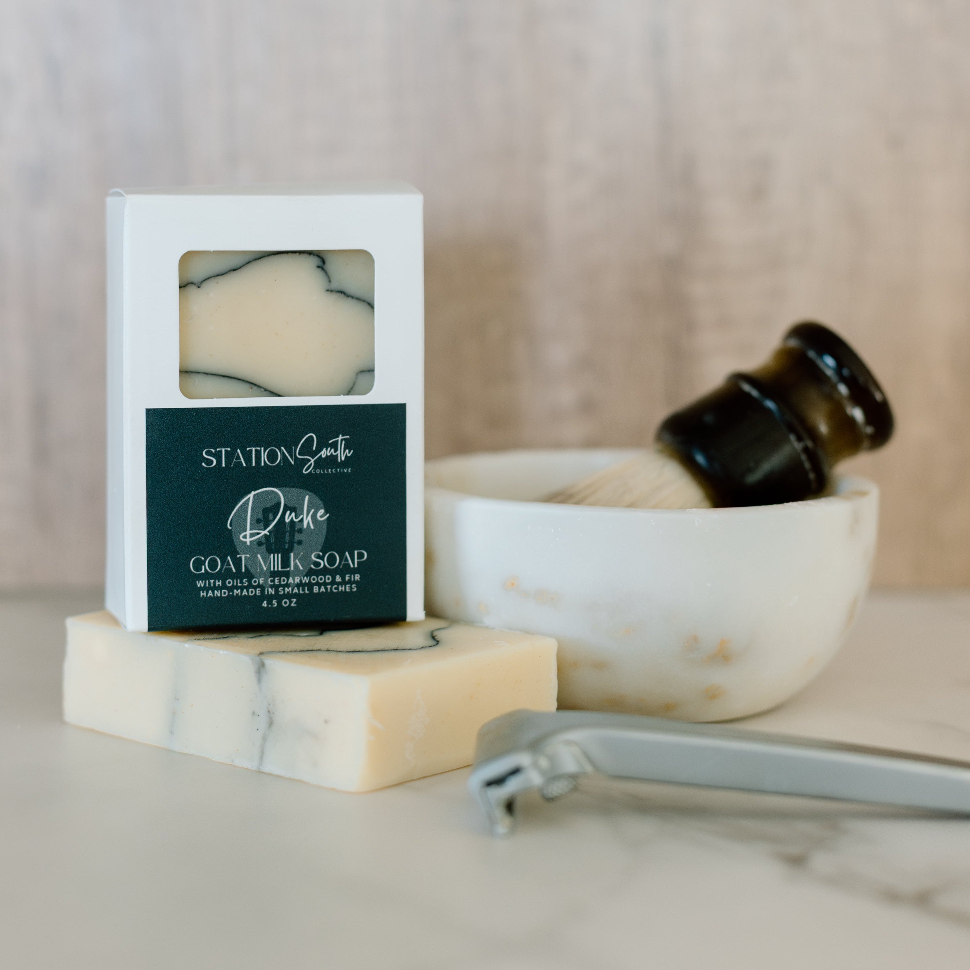 Bar of soap in a white box with a label, next to a bowl and shaving tools on a marble surface.