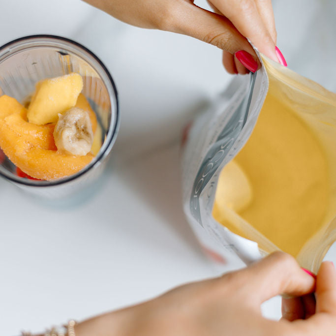 A top-down view of a person pouring a thick yellow powder from a resealable pouch into a blender cup containing frozen fruit, on a light marble surface.
